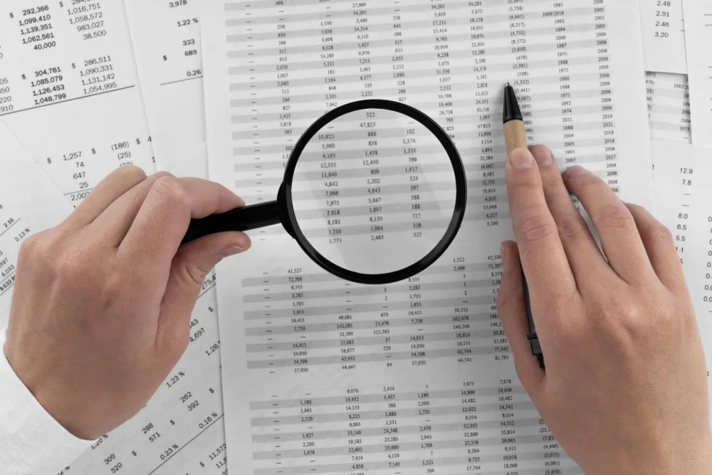 Somebody holding a magnifying glass to paperwork covered in numbers.